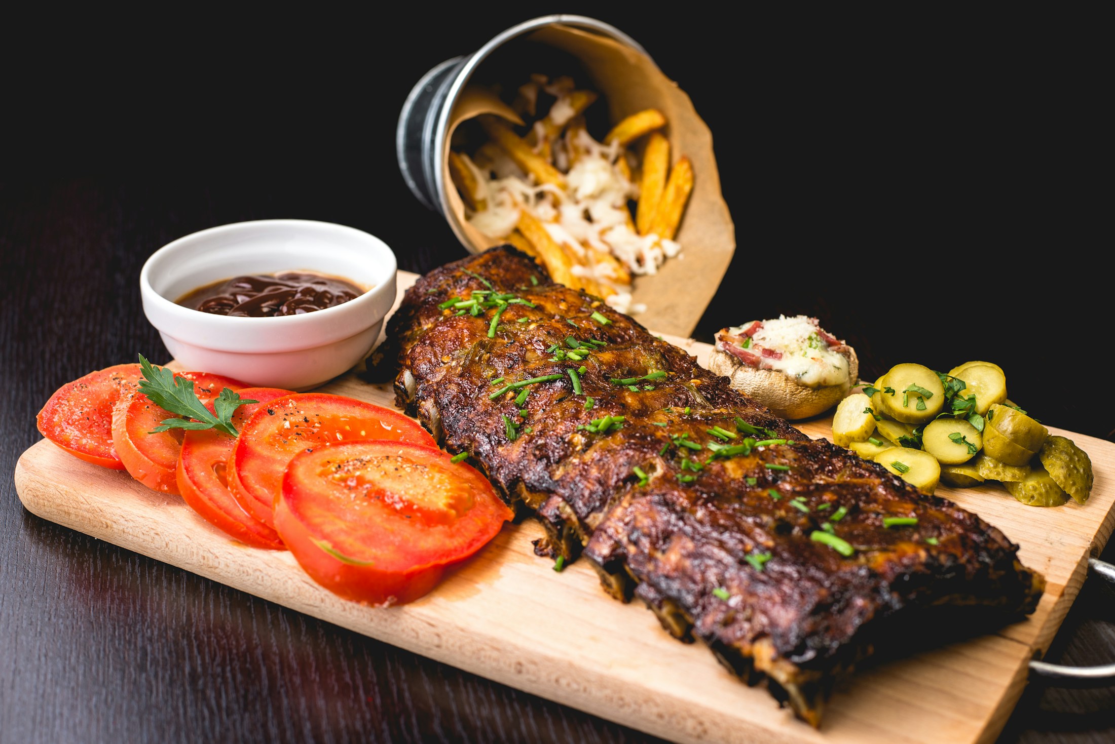 A wood board of glistening Brazilian-style barbecue ribs with tomatoes, fries and pickles, warmly lit against a dark table